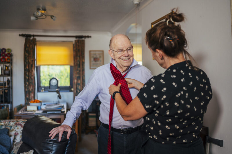 A woman helping a man put on a tie. The man is smiling