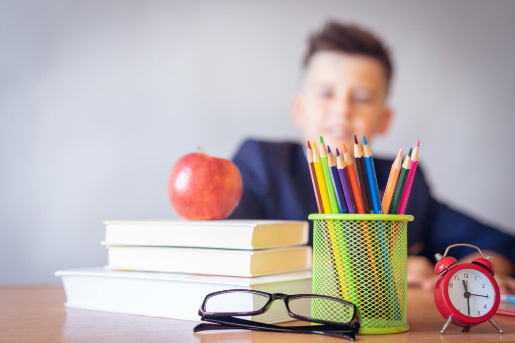 A stack of books with an apple on top alongside a container of coloured pencils, a clock and a pair of glasses. There is also a blurred image of a child in the background.